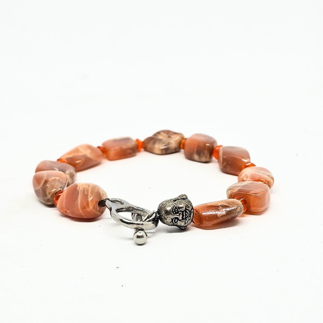 A Sunstone bracelet with Carnelian Spacers and a Buddha Charm, featuring a series of round beads in shades of orange and red, with a silver-colored Buddha charm and a closure mechanism.